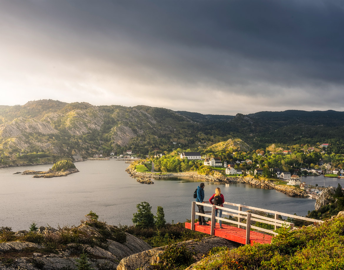Two people standing outdoors and appreciating the view of rocky cliffs, calm water, and quaint homes in Brigus
