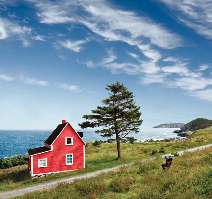 Man sits restfully in an open field, taking in the view of a beautiful red house by the water in Tors Cove