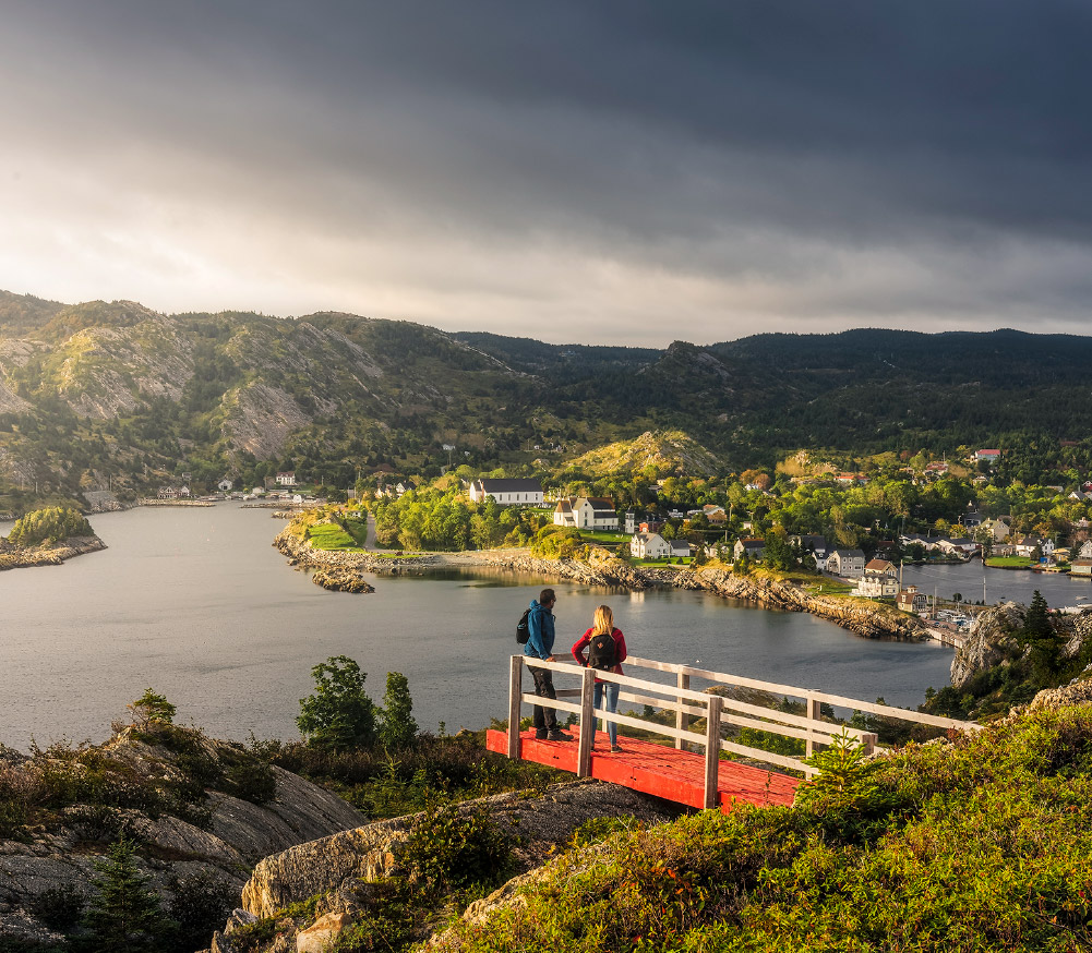Two people standing outdoors and appreciating the view of rocky cliffs, calm water, and quaint homes in Brigus