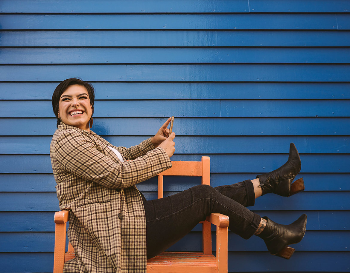Woman smiles on a chair while holding her phone in front of a blue clapboard house