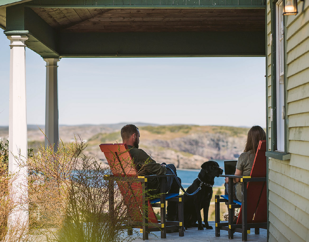 Man and woman work on their patio, overlooking a gorgeous view of seaside cliffs in Trinity as their dog sits by them