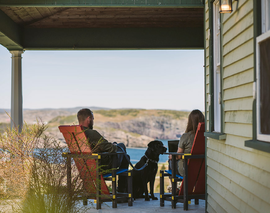 Man and woman work on their patio, overlooking a gorgeous view of seaside cliffs in Trinity as their dog sits by them