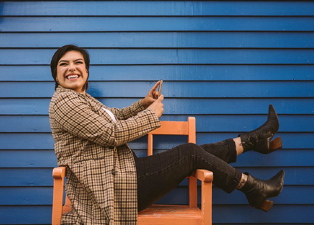 Woman smiles on a chair while holding her phone in front of a blue clapboard house