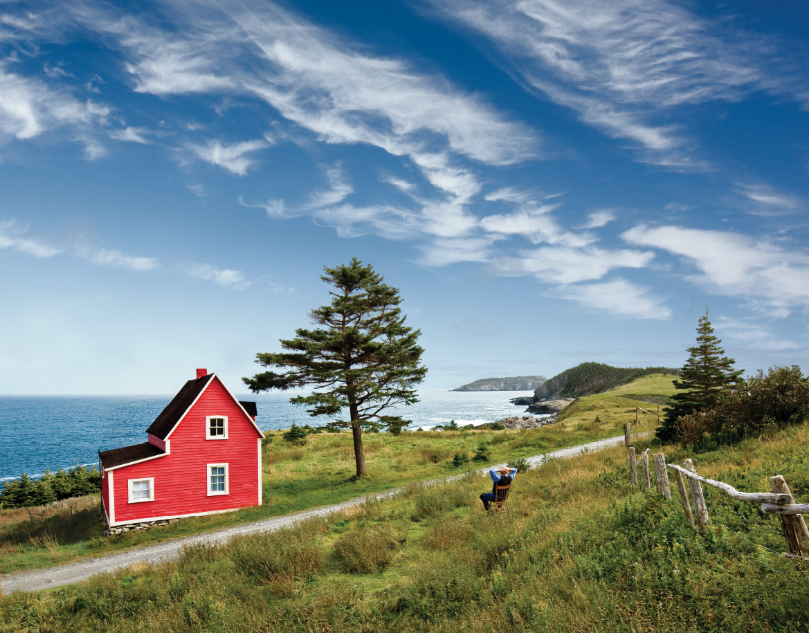 Man sits restfully in an open field, taking in the view of a beautiful red house by the water in Tors Cove